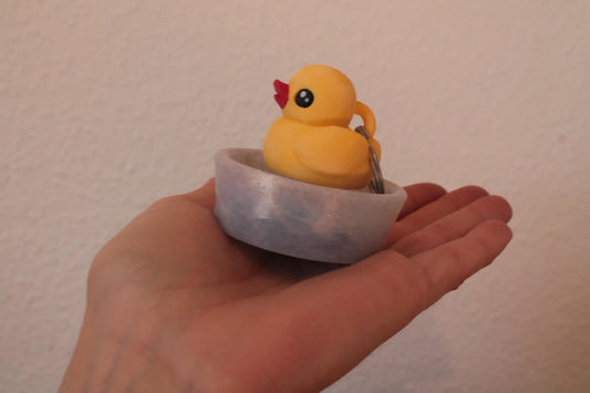 Hand holding a small yellow rubber duck keychain in a round marbled dish