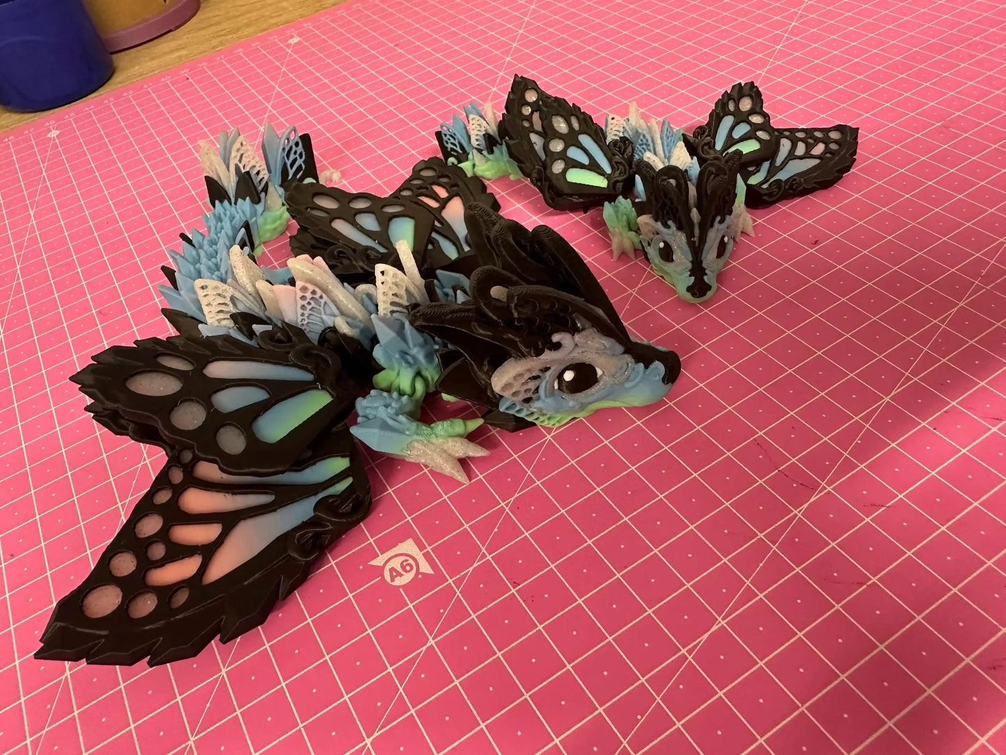 3D printed butterfly dragon figurines with pastel wings on a pink grid cutting mat