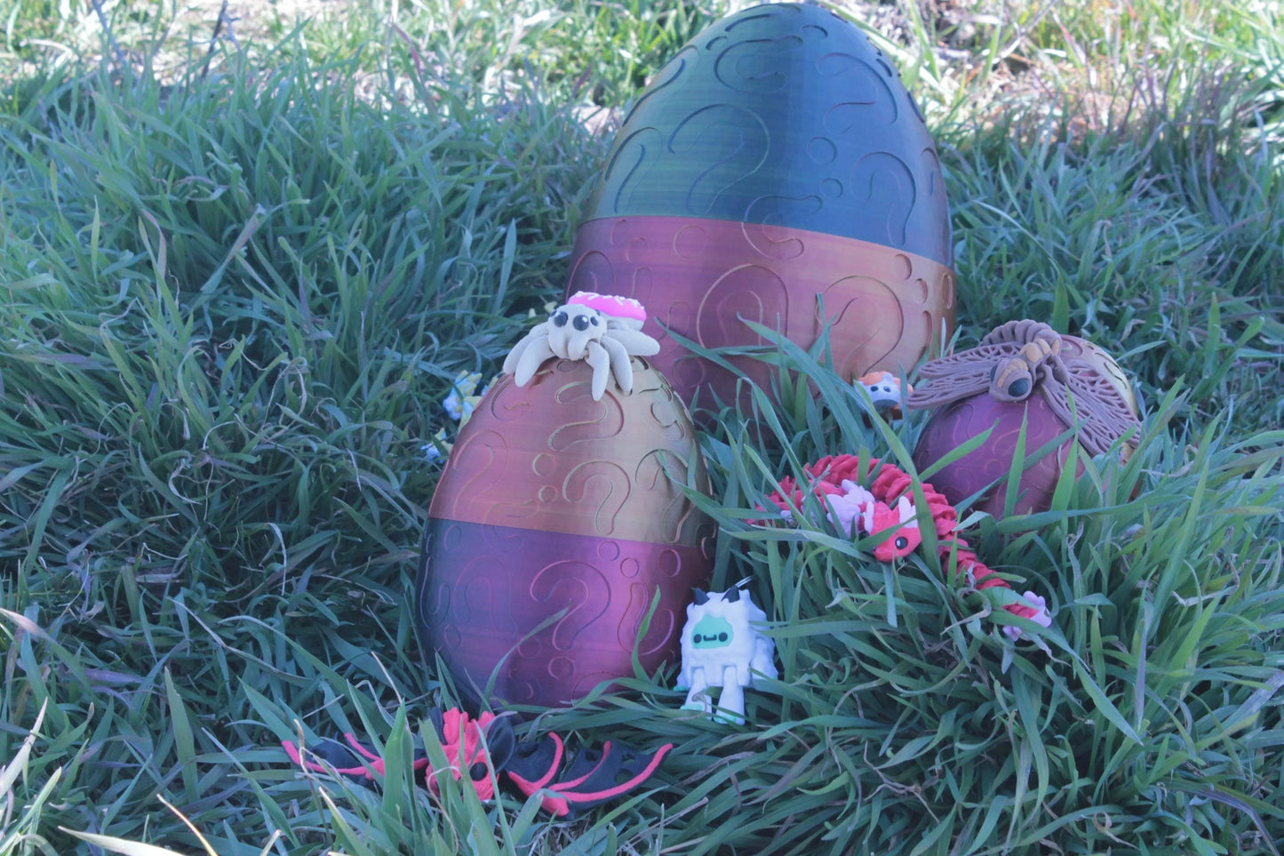 Überraschungs-Ei mit Figuren: großes violett-blaues Ei und zwei kleinere im Gras, pinke Deko