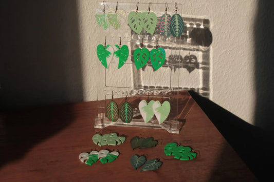 Assorted green acrylic leaf earrings on a clear display stand, with sunlight casting shadows.